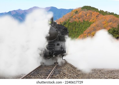 A steam locomotive emitting white smoke with a backdrop of mountains filled with autumn leaves