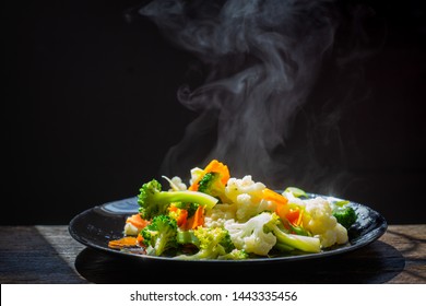 The Steam From The Vegetables Carrot Broccoli Cauliflower In A Black Plate  , A Steaming. Boiled Hot Healthy Food On Table On Black Background,hot Food And Healthy Meal Concept