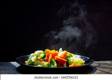 The Steam From The Vegetables Carrot Broccoli Cauliflower In A Black Plate  , A Steaming. Boiled Hot Healthy Food On Table On Black Background,hot Food And Healthy Meal Concept