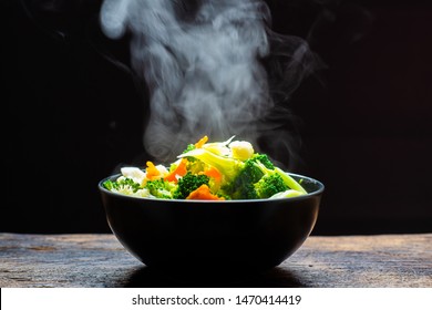 The Steam From The Vegetables Carrot Broccoli Cauliflower In A Black Bowl , A Steaming. Boiled Hot Healthy Food On Table On Black Background,hot Food And Healthy Meal Concept