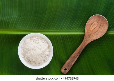 Steamed Cook Jasmine White Rice In Bowl With Wooden Spoon On Green Banana Leaf. Top View.