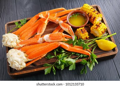 Steamed Crab Legs With Melted Butter, Garlic Cloves, Lemon Slices, Grilled Corn In Cobs And Fresh Parsley On Old Wooden Cutting Board, On Black Wooden Table, View From Above