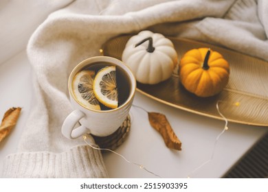 A steaming cup of tea with slices lemon rests on a woven coaster beside a small decorative pumpkin, with soft knit blankets and treats nearby, all illuminated by warm sunlight. Cozy autumn concept. - Powered by Shutterstock