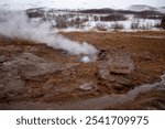 Steaming geothermal vent on a barren, snow-dusted terrain with overcast skies.