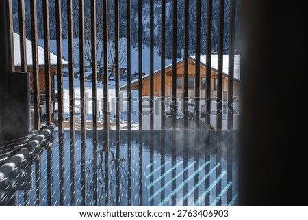 Steamy indoor spa pool frames a snow covered Dolomites village in Italy. Chalet roofs and fir lined slopes sit beyond. Midday sun casts striped reflections on water.