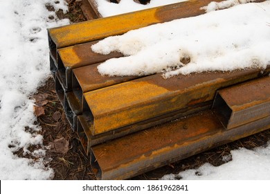Steel beams stored outdoors are covered with rust, wet snow lies on them