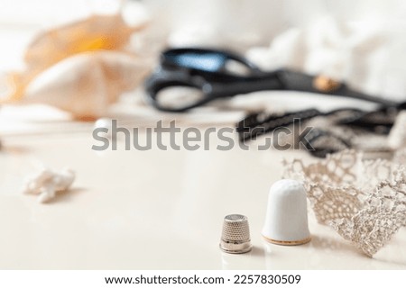 A steel and ceramic thimble stand on a white background. To the right is lace. Blurred scissors in the background

