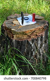 Steel Sharp Blade Of Folding Hunting Knife Stuck In Old Cut Tree Stump In The Wild Forest And Lying On It Metal Flashlight, Compass, Magnifying Glass And Vintage Military Map Vertical View