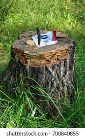 Steel Sharp Blade Of Folding Hunting Knife Stuck In Old Cut Tree Stump In The Wild Forest And Pressing Vintage Military Map Metal Flashlight, Compass, Magnifying Glass Full View