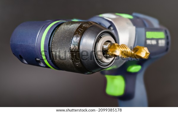 Steel tap and drill bit in power screwdriver right\
angle head on a gray background. Closeup of spiral fluted cutting\
tool with gold titanium coating clamped in jaws of cordless\
drilling machine chuck.