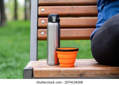 Steel thermos and silicone mug. Tourist dishes on the bench. Drinking coffee in the park.