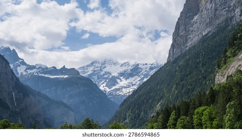Steep alpine cliffs rise dramatically above a lush green valley, framing distant snow-covered peaks beneath drifting clouds capturing the untouched beauty of the Swiss Alps.