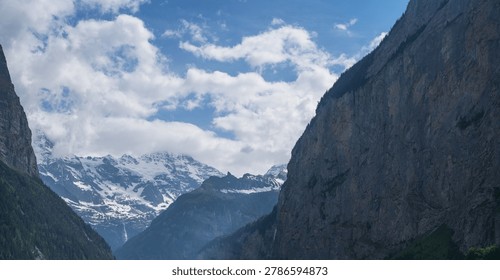 Steep alpine cliffs rise dramatically above a lush green valley, framing distant snow-covered peaks beneath drifting clouds capturing the untouched beauty of the Swiss Alps.