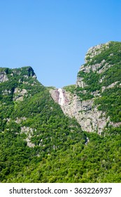 Steep cliffs covered in vegetation with distant waterfall flowing from top of mountain under clear blue sky, at Western Brook Pond, Gros Morne National Park, Newfoundland, Canada.
