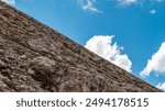 A steep rocky slope meets the vibrant blue sky, dotted with fluffy white clouds