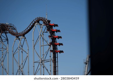 A steep roller coaster drop supported by tall metal beams, with red coaster cars descending the track under a clear blue sky. The dramatic angle and visible riders highlight the thrilling motion and engineering complexity of the attraction.