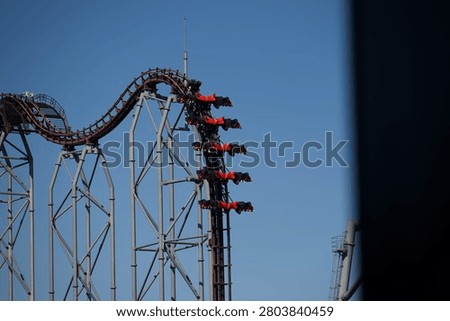 A steep roller coaster drop supported by tall metal beams, with red coaster cars descending the track under a clear blue sky. The dramatic angle and visible riders highlight the thrilling motion and engineering complexity of the attraction.