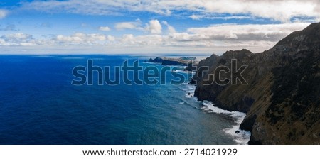 Steep volcanic cliffs meet deep blue water near Ponta de Sao Lourenco, Madeira, Portugal, daylight with broken clouds, white surf lines the shore, vast sea and rock forms.