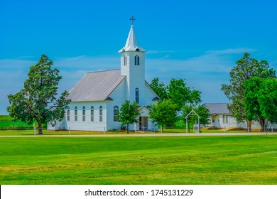 A steepled white church sits in a spring  meadow in Texas. Trees surround the Christian chapel. 