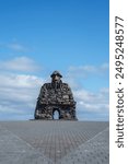 The Stekkjarkot stone sculpture, resembling a human figure with an archway, stands on a paved platform in Iceland. Clear blue sky and scattered clouds enhance its rugged design.