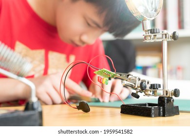 STEM Education And 21st Century Skills Concept. Young Asian Teenager, Kid Is Cutting Wire For Water Moisture Sensor Circuit Board On Grip Hand Desktop Stand. DIY School Science And Technology Project.