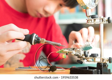 STEM Education And 21st Century Skills Concept. Young Asian Teenager, Kid Is Soldering Water Moisture Sensor Circuit Board With Grip Hand Desktop Stand. DIY School Science And Technology Project.