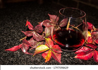 A Stemless Glass Filled With A Dark Red Wine, Surrounded By Autumn Leaves In Different Shades Of Red On A Black Countertop