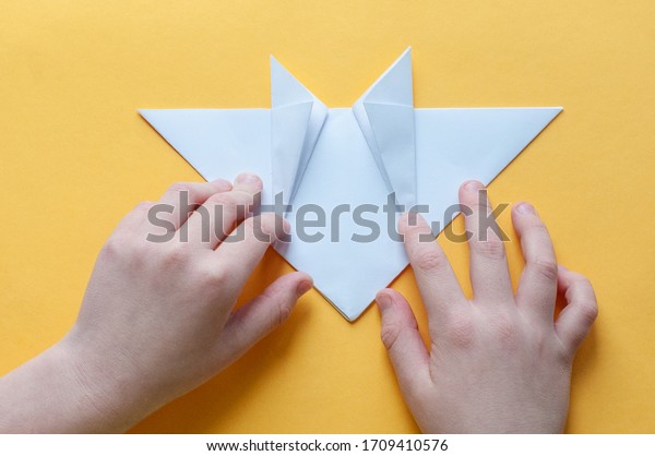 Step\
12. Hands of a child on a yellow background divide a square of\
white paper into triangles. Origami bull\
concept.