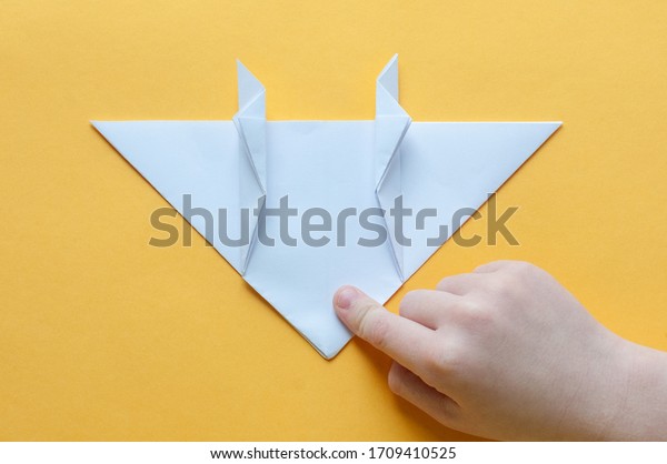 Step\
14. Hands of a child on a yellow background divide a square of\
white paper into triangles. Origami bull\
concept.
