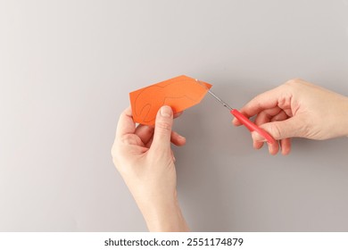 step in creative project, top down view one hand hold detail from carton and another hand hold red handle scissors which cutting out , claws made from orange paper, cut out details, grey background,