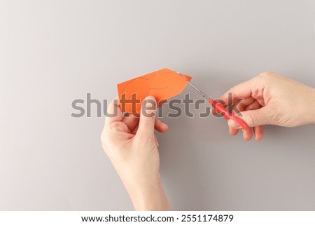 step in creative project, top down view one hand hold detail from carton and another hand hold red handle scissors which cutting out , claws made from orange paper, cut out details, grey background,