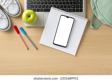 Step into the world of online studying from home with this captivating top-down view, presenting laptop, smartphone, rucksack, shoes, notepad, pens and apple on wooden background - Powered by Shutterstock