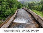 Stepped auxiliary spillway with water flowing in Robertville Dam towards Warche River in valley, green trees and wild vegetation in misty and blurry background, cloudy day in Waimes, Belgium