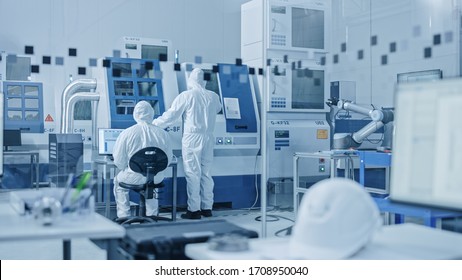 Sterile Modern Factory: Professionals In Coveralls, Masks Working On CNC Machinery. Medical Electronics Manufacturing Laboratory With High Tech Robot Arm Production Line And Contemporary Equipment