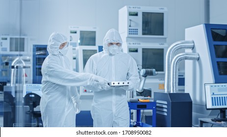 Sterile Modern Factory: Professionals In Coveralls, Masks Walk Through Workshop And Talk. Medical Electronics Manufacturing Laboratory With High Tech CNC Machinery, Robot Arm Production Line.