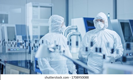 Sterile Modern Factory: Professionals In Coveralls, Masks Walk Through Workshop And Talk. Medical Electronics Manufacturing Laboratory With High Tech CNC Machinery, Robot Arm Production Line Equipment