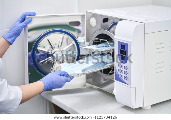Sterilizing medical instruments in autoclave.\
Dental office. Dentist assistant\'s hands get out sterilizing\
medical instruments from autoclave. Selective focus. Sterile dental\
tools\
