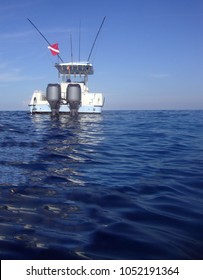 Stern Or Back Of A Baby Blue Sport Fisher Boat With Out Riggers And Down Rigger In The Open Ocean Flying A Red And White Diving Flag Against A Deep Blue Sea And Light Blue Sky.