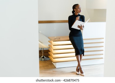 Stern Elegant Business Woman Wearing Black Dress And Beige Shoes In Light Office Looking Towards With Her Agenda, Full Length Portrait