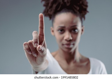 Stern Focused Young Woman Holding Up Her Index Finger As Though Emphasising A Point, Calling A Halt, Or Identifying Herself On A Grey Studio Background