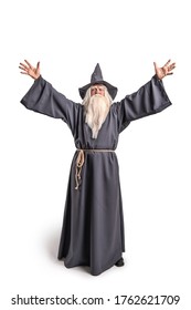 A stern grey-haired bearded wizard in a gray cassock and a cap is practicing sorcery and doing magic against a white background. - Shutterstock ID 1762621709