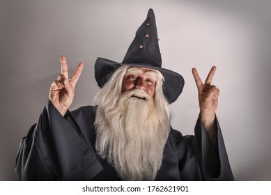 A stern grey-haired bearded wizard in a gray cassock and a cap is practicing sorcery and doing magic against a white background. - Shutterstock ID 1762621901