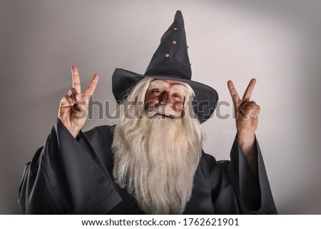 A stern grey-haired bearded wizard in a gray cassock and a cap is practicing sorcery and doing magic against a white background.