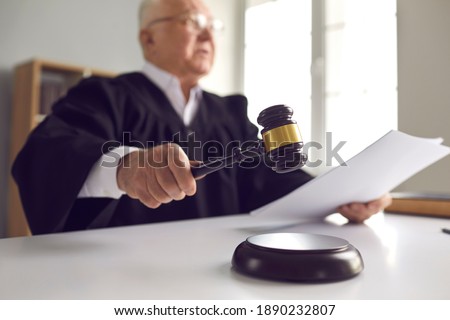 Stern judge with paper document pronouncing sentence in a court of law. Judge finds the accused guilty, passes judgement and rules case closed. Hand holding gavel and hitting sound block in close-up