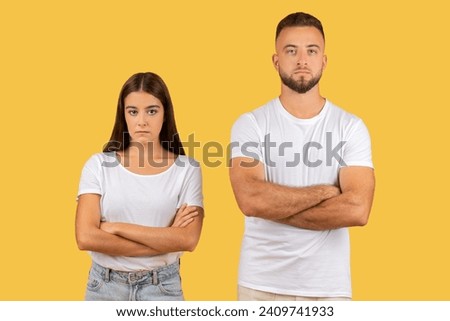 Stern-faced young european couple with arms crossed in white t-shirts, exuding a strong sense of disagreement or defiance, standing against a uniform yellow background, studio