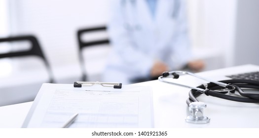 Stethoscope, Clipboard With Medical Form Lying On Hospital Reception Desk With Laptop Computer And Busy Doctor And Patient Communicating At The Background. Medical Tools At Doctor Working Table