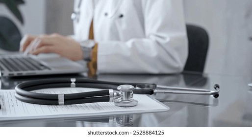 Stethoscope and clipboard with medication history records are on a glass desk behind a physician using a laptop computer. Medicine concept - Powered by Shutterstock