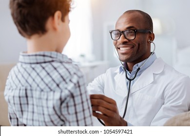 Stethoscope exam. Attractive cheerful male doctor listening to boy while putting on glasses and using stethoscope