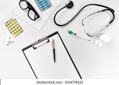 Stethoscope In The Office Of Doctors.Top View Of Doctor's Desk Table, Blank Paper On Clipboard With Pen. Copy Space. Designer's Blank