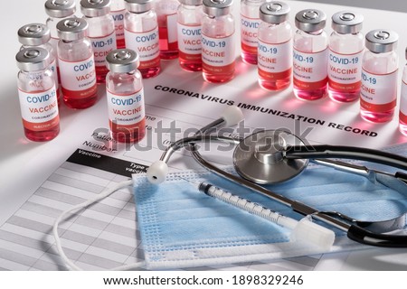 Stethoscope, syringe and vaccine vial glass bottles for vaccination against COVID-19 SARS-CoV-2 coronavirus pandemic lying on surgical masks and on the immunization document. 